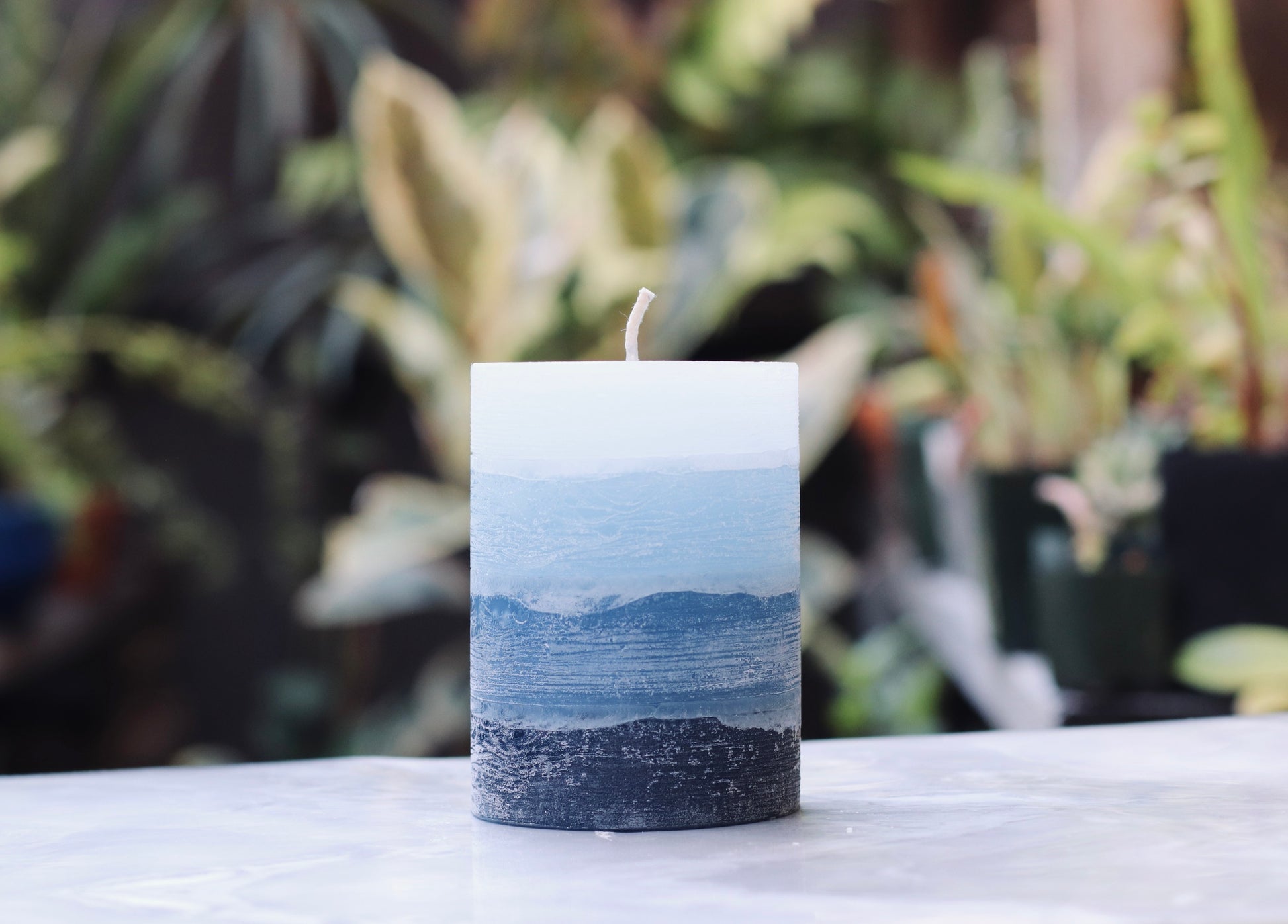 Blue rustic candle on a marble surface with plants in the background
