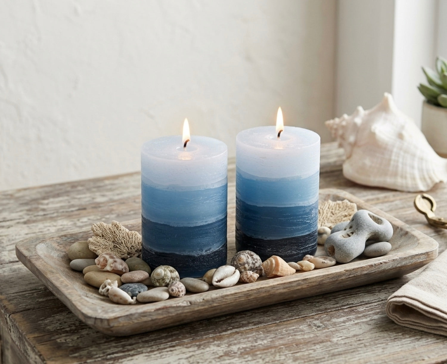 Two blue gradient candles on a wooden tray with pebbles and shells.