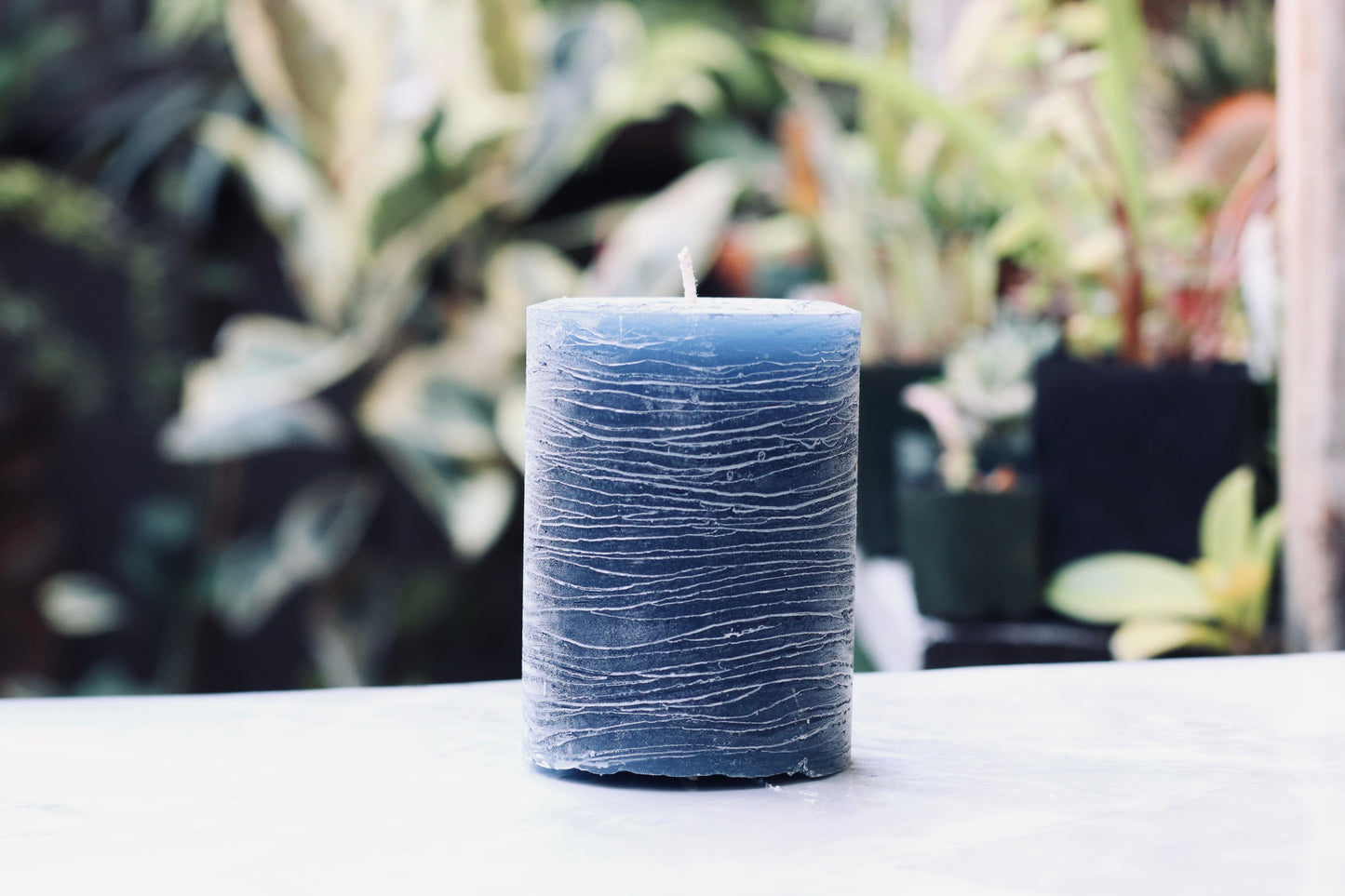 Navy blue textured candle on a white surface with a blurred plant background