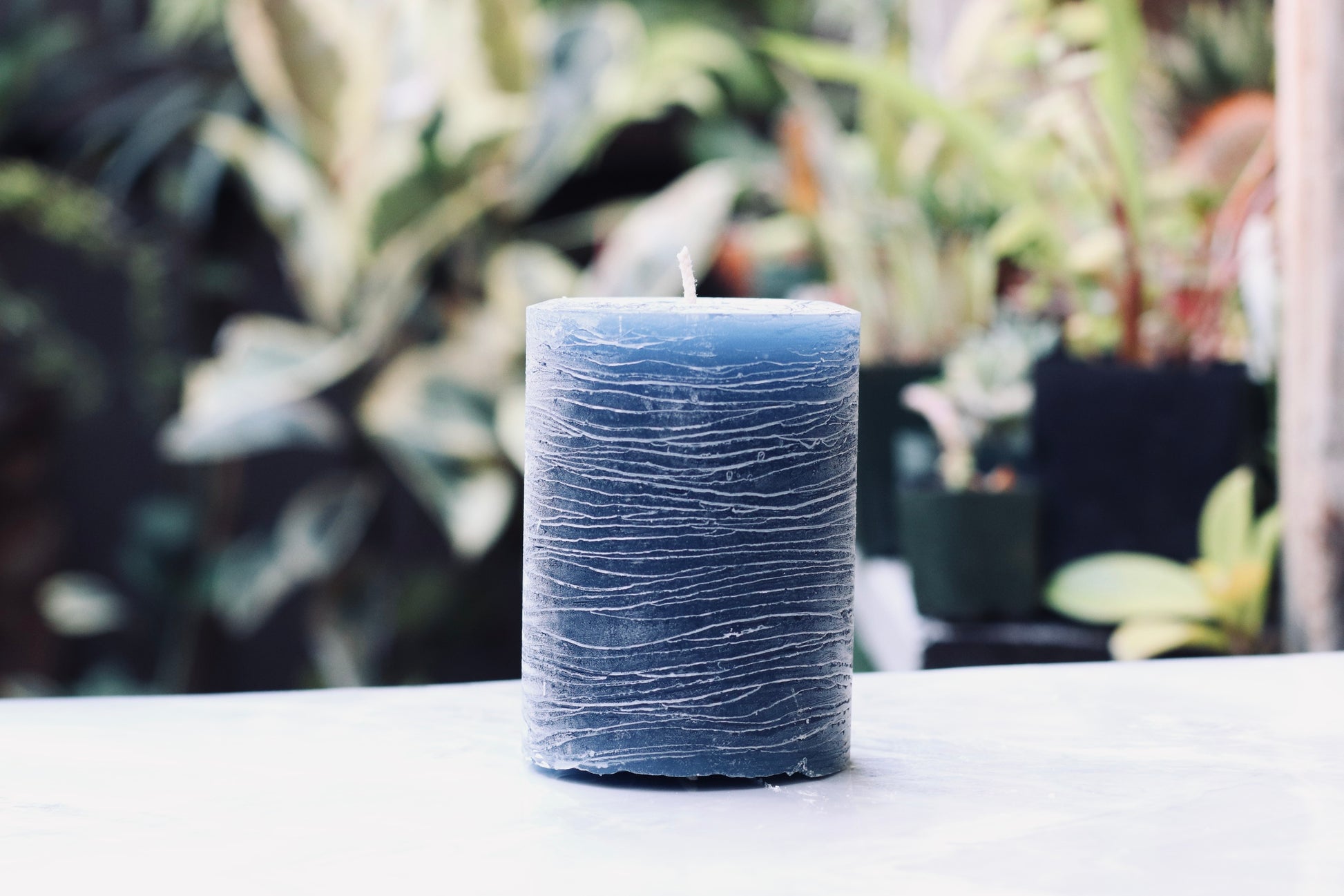 Navy blue textured candle on a white surface with a blurred plant background