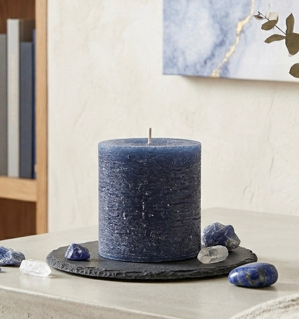 A navy blue candle on a black stone coaster with decorative stones and a painting in the background.
